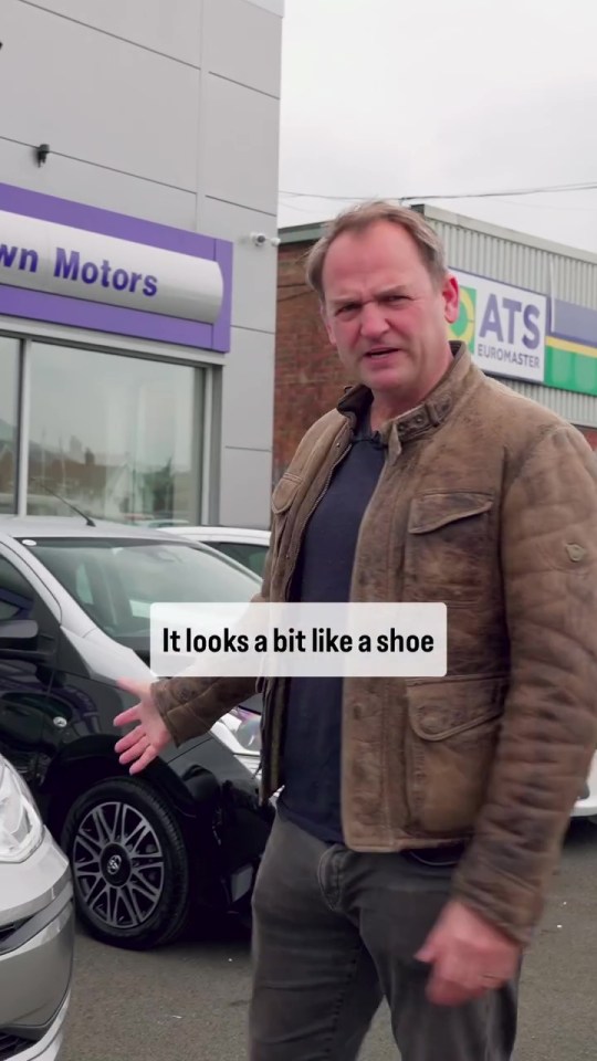 Man in brown jacket standing in front of cars, saying a car looks like a shoe.