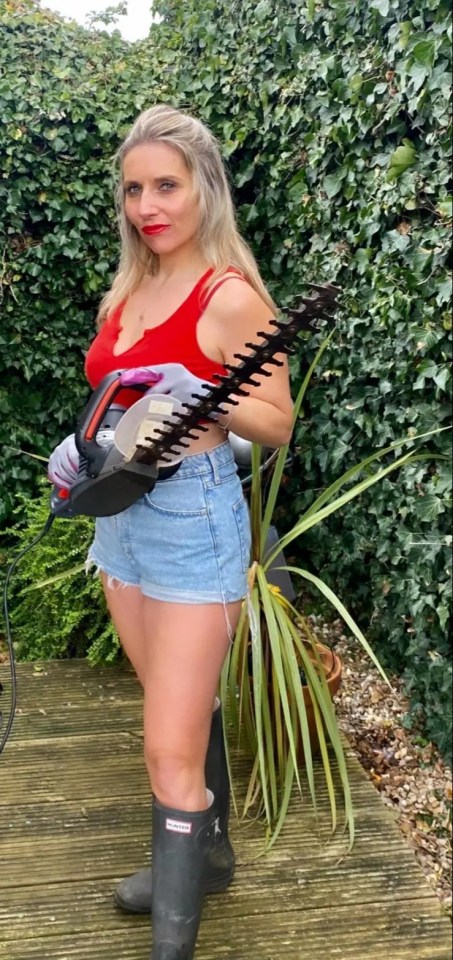 Woman in red top and denim shorts using a hedge trimmer in her garden.
