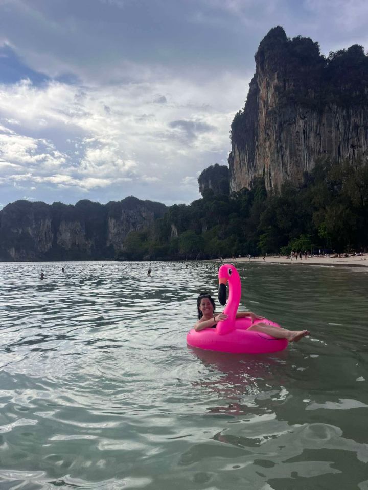 Woman on pink flamingo float in calm water with cliffs in the background.