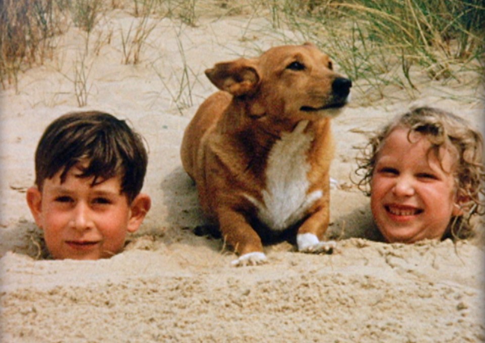 Young Prince Charles and Princess Anne buried up to their necks in sand with a dog.