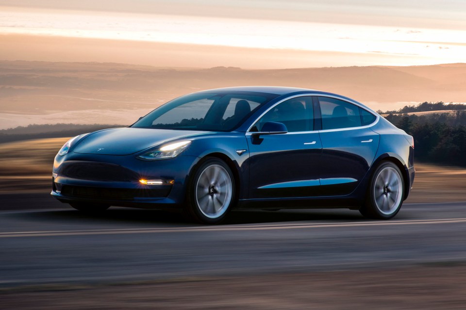 Tesla Model 3 driving on a road at sunset.