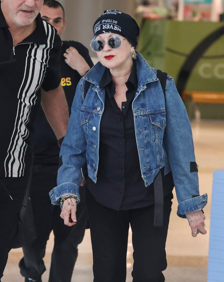 Cyndi Lauper at an airport wearing a denim jacket and hat.