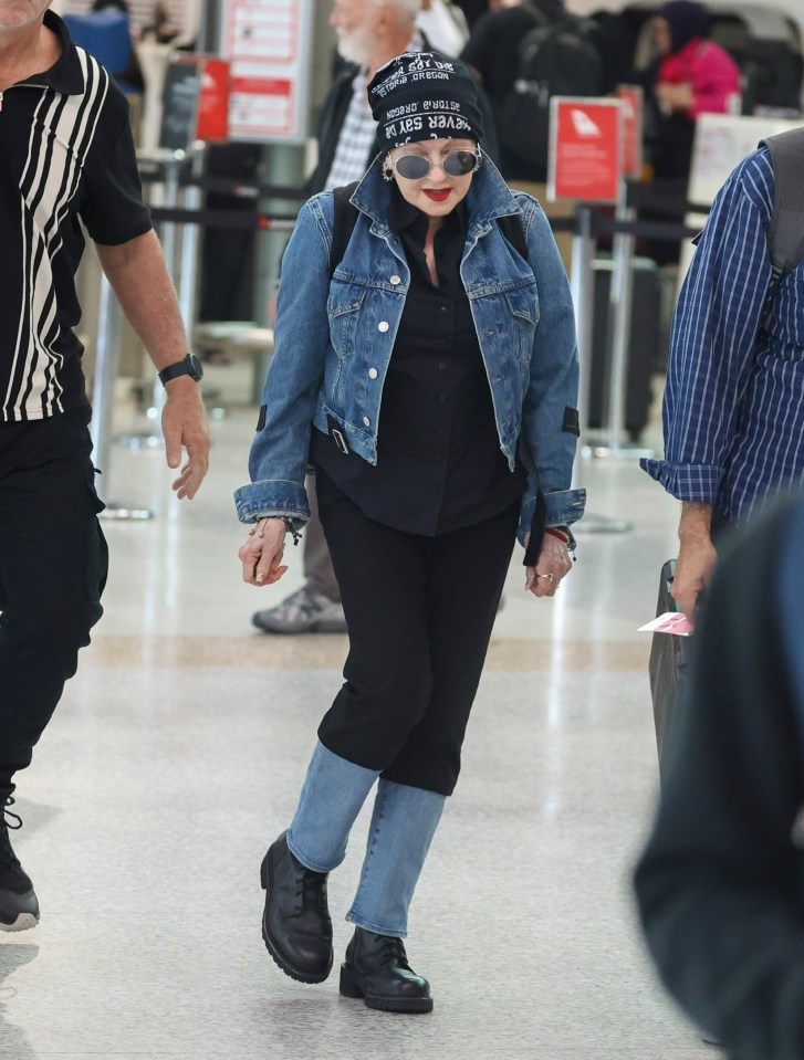 Cyndi Lauper at an airport wearing a denim jacket and hat.