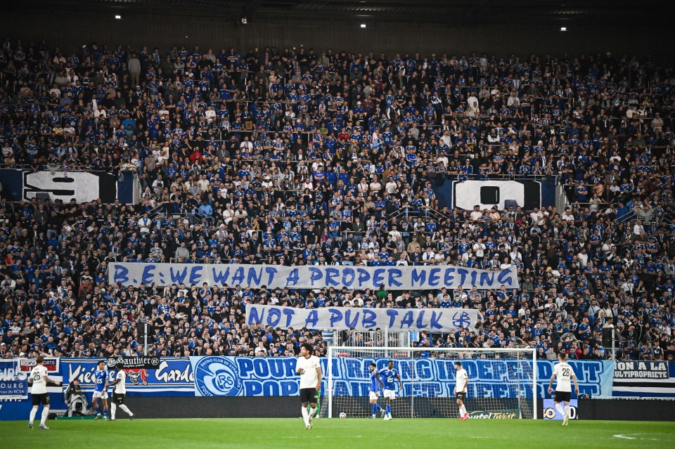 Stadium crowd holding a banner that reads "B.E. WE WANT A PROPER MEETING NOT A PUB TALK".