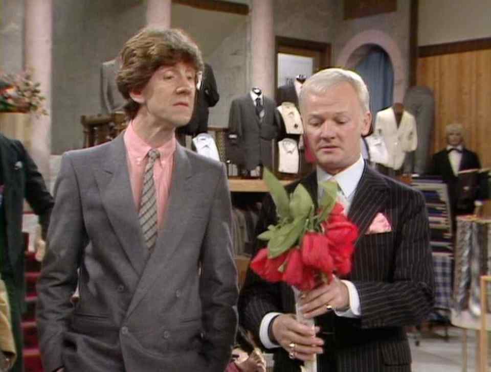 Two men in suits in a menswear shop; one holds a bouquet of red flowers.