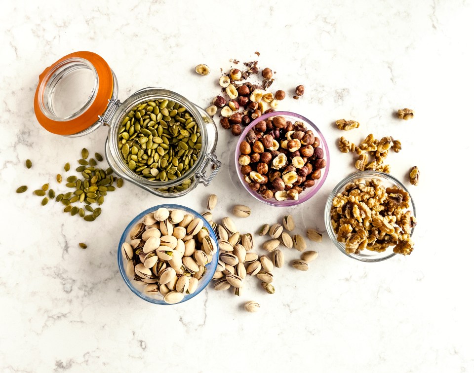 Overhead view of pistachios, hazelnuts, walnuts, and pumpkin seeds in bowls and a jar.