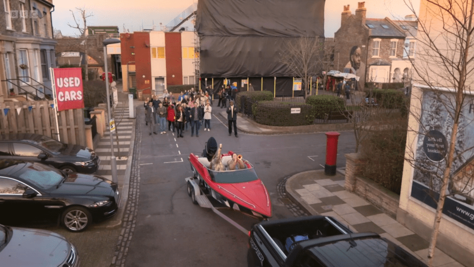 A red speedboat being towed down a residential street, with a crowd of people watching.