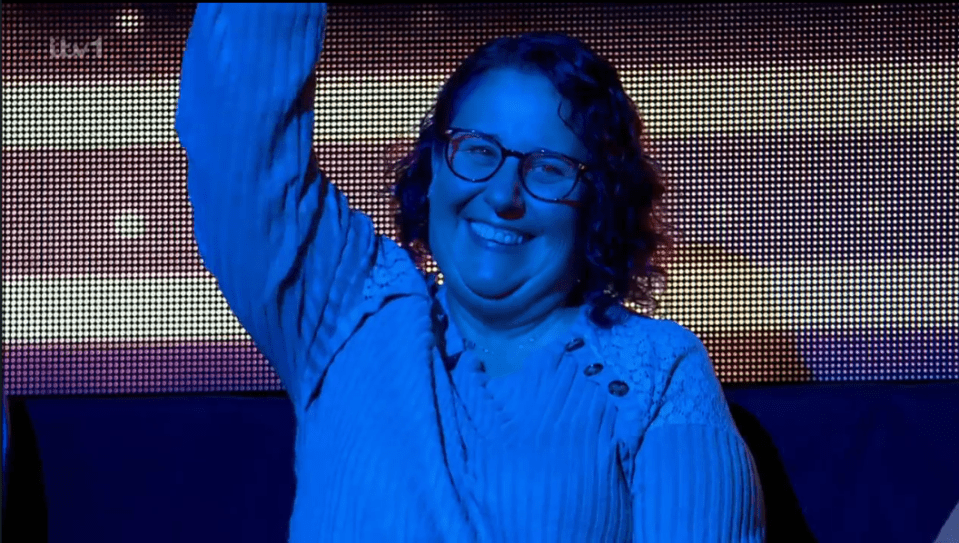 Screenshot of a woman wearing glasses, smiling and raising her hand.