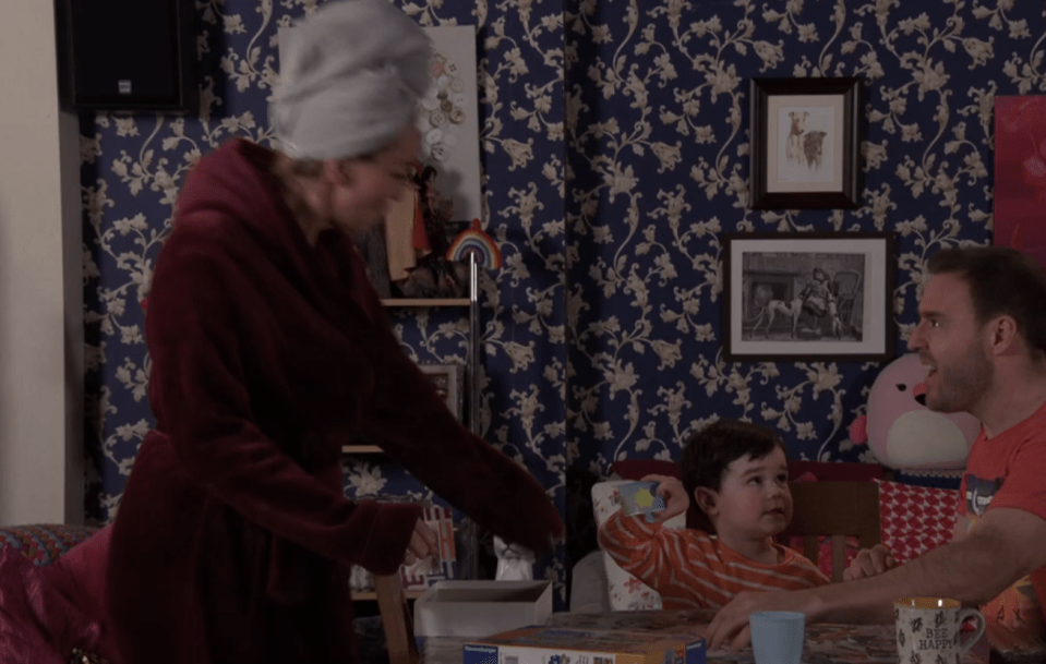 Screenshot of a woman in a robe interacting with a man and child at a table.