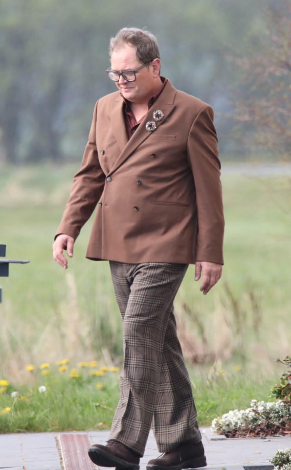Alan Carr walking outside wearing a brown suit and plaid pants.