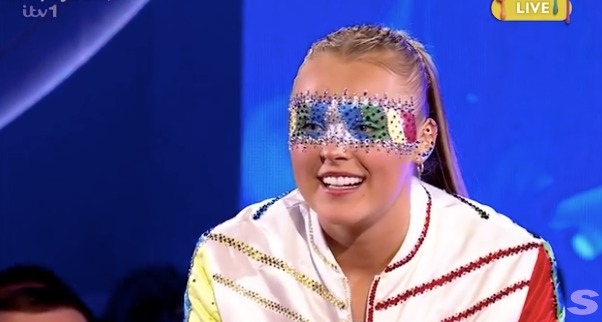 Screenshot of a woman wearing a colorful jeweled mask on a game show.