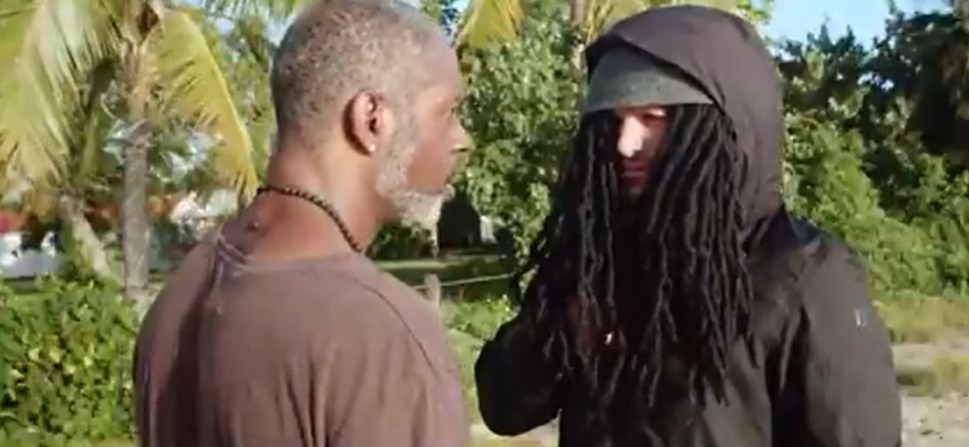 Still image from a movie showing two men talking; one man has dreadlocks.