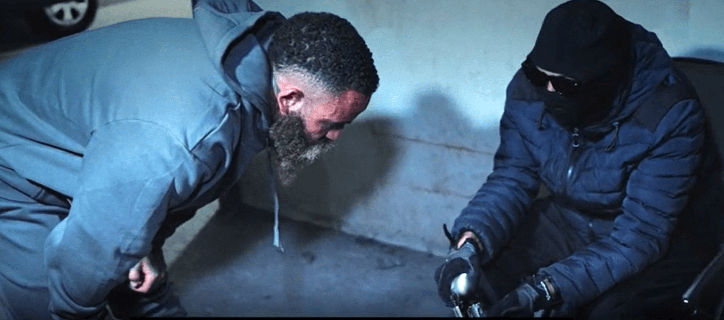 Ashley Cain bending down to look at something held by a person wearing a balaclava.