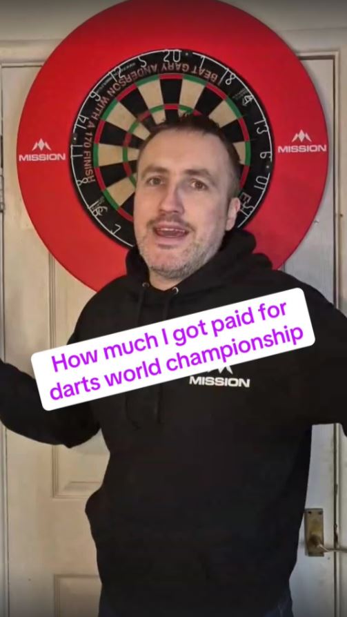 Man in front of a dartboard discussing his darts world championship earnings.
