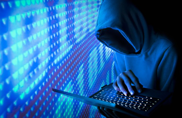 Hooded figure using a laptop in front of a blue screen.