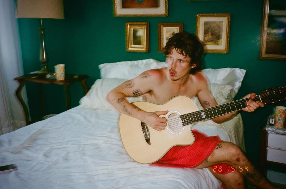 A shirtless man sitting on a bed playing an acoustic guitar.