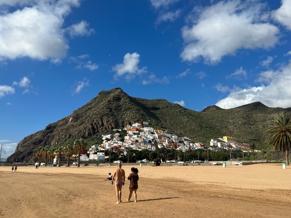 Couple walking on a sandy beach with a town on a hillside in the background.