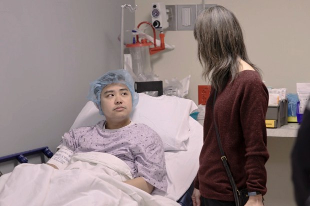 A man in a hospital gown sits in a hospital bed talking to a woman.