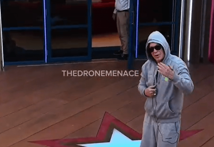 Man in gray hoodie and sunglasses gesturing.