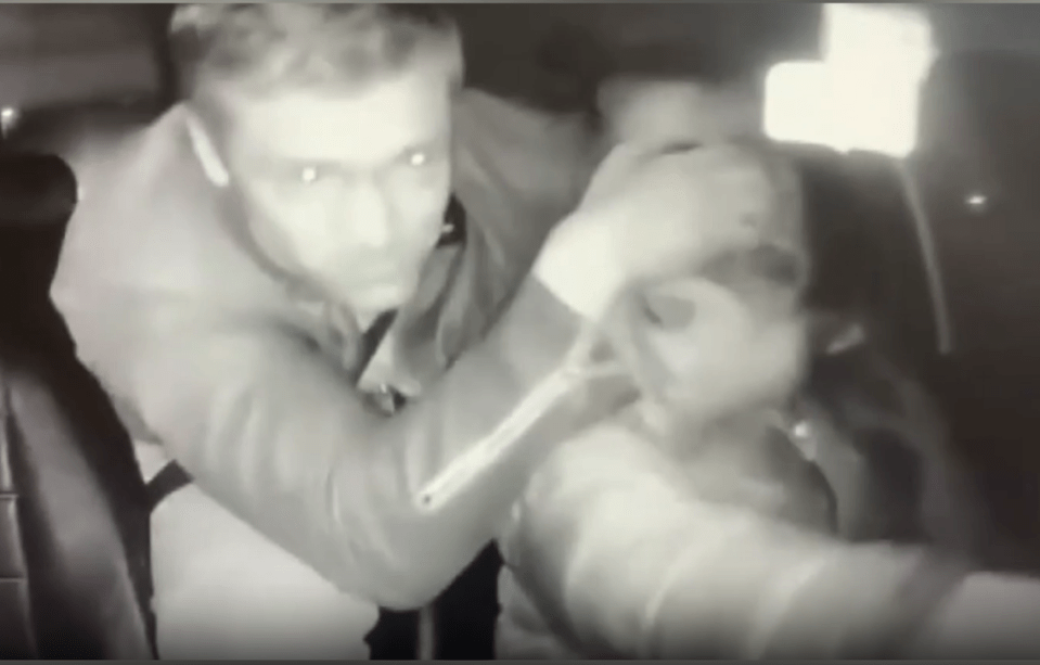 Grainy black and white security camera footage of a man assaulting a person in a vehicle.