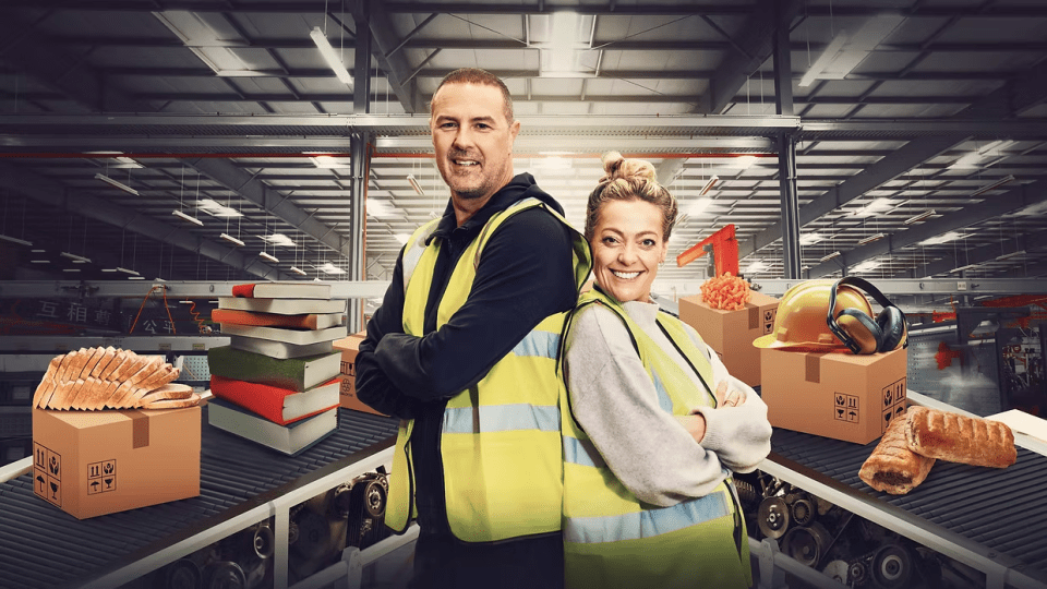 Two people in high-visibility vests stand back-to-back in a factory setting with conveyor belts carrying books, bread, and sausage rolls.