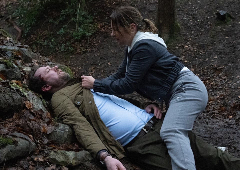 A woman kneeling over an injured man in a wooded area.