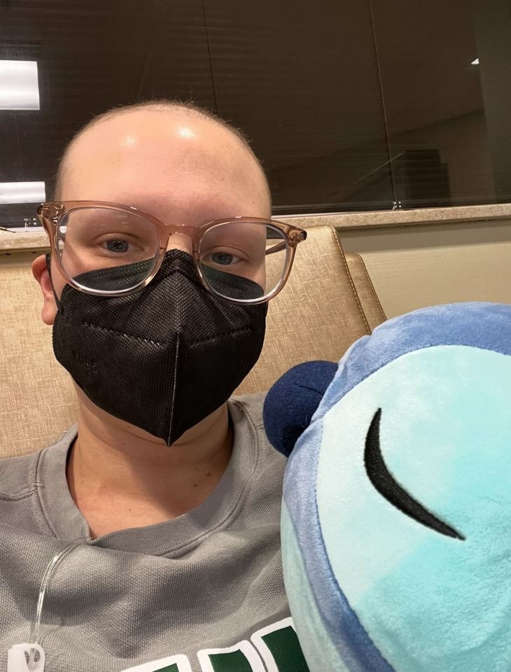 A person with glasses and a face mask sits in a chair holding a plush toy.