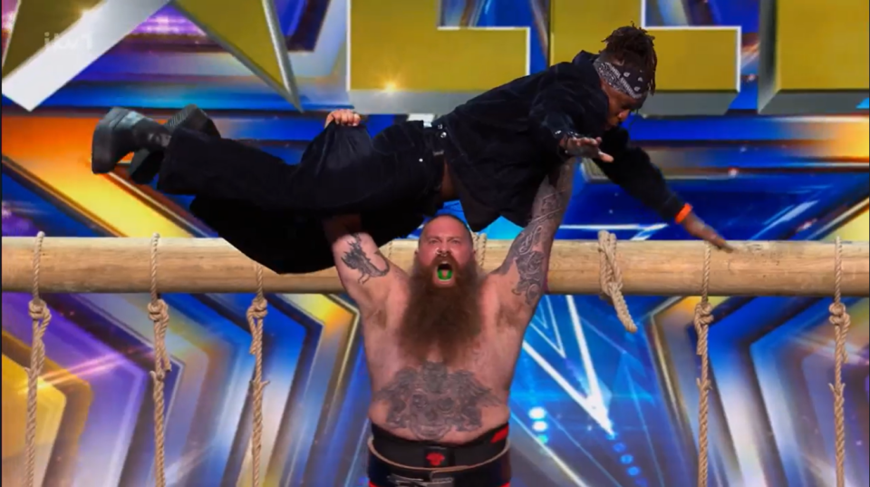 A strongman lifting another man over a wooden beam.