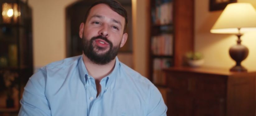 A man in a light blue shirt speaks to the camera.