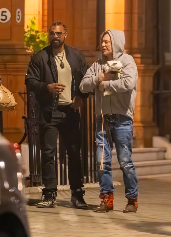 Mickey Rourke and another man leaving a London hotel at night, with Rourke holding a small dog.