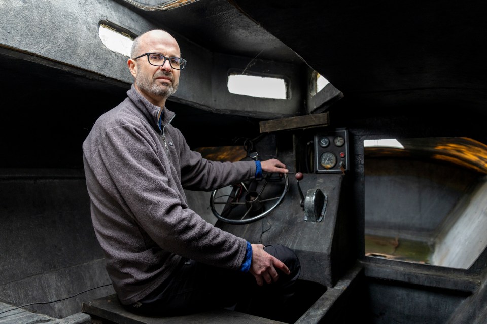 Man sitting at the controls of a small, dark submarine.