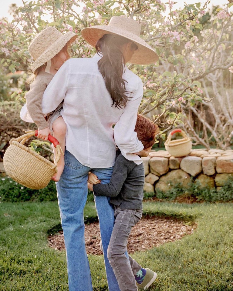 Woman carrying a young child, with another child holding onto her leg, in a garden.