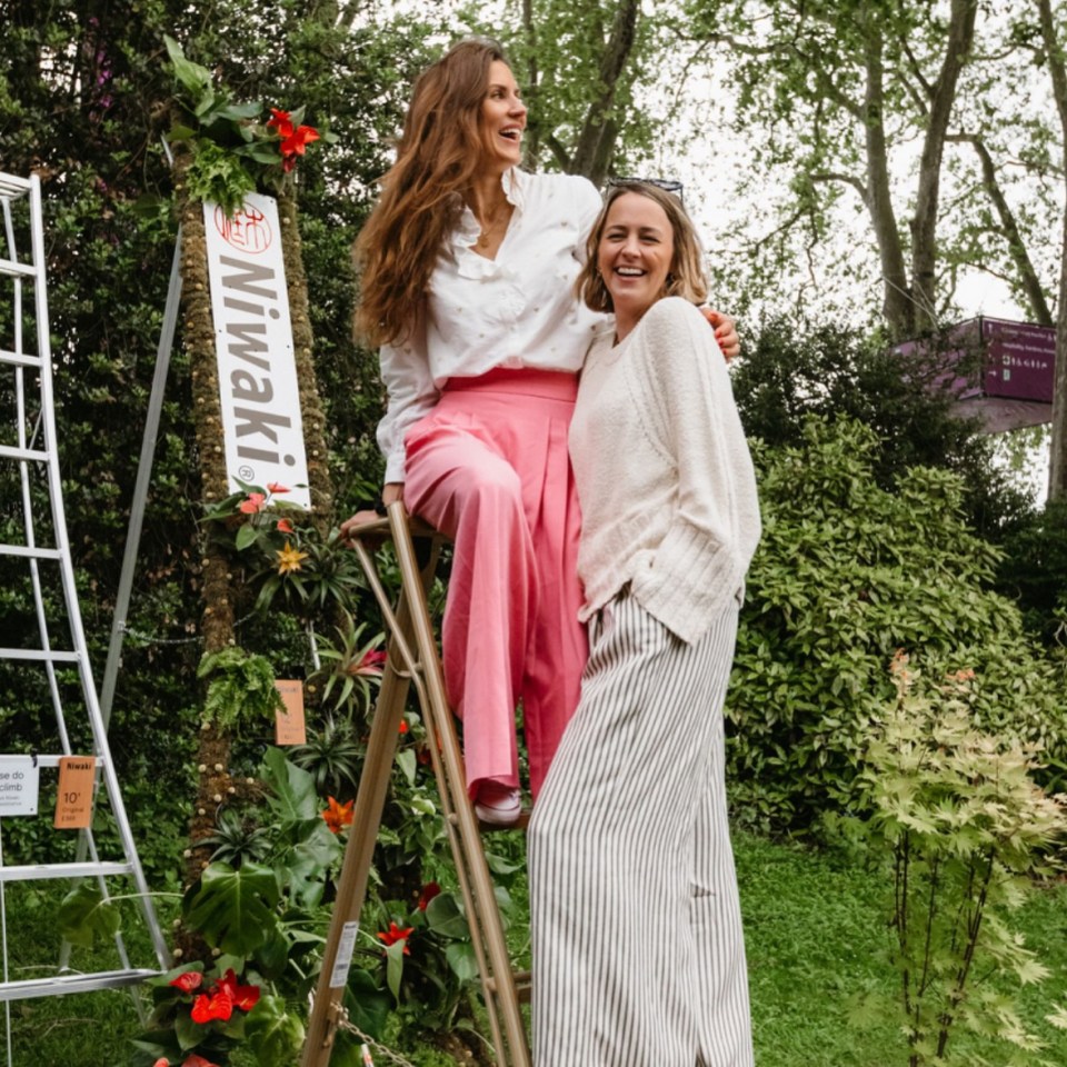 Two women smiling, one sitting on a stepladder in front of a vertical garden display.