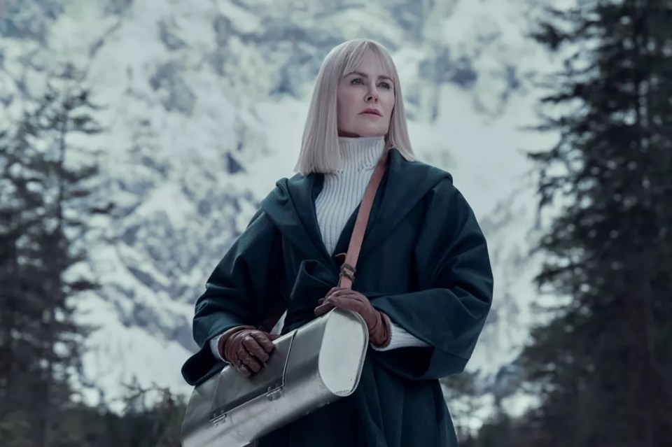 Woman in a green coat carrying a metal case in a snowy mountain landscape.