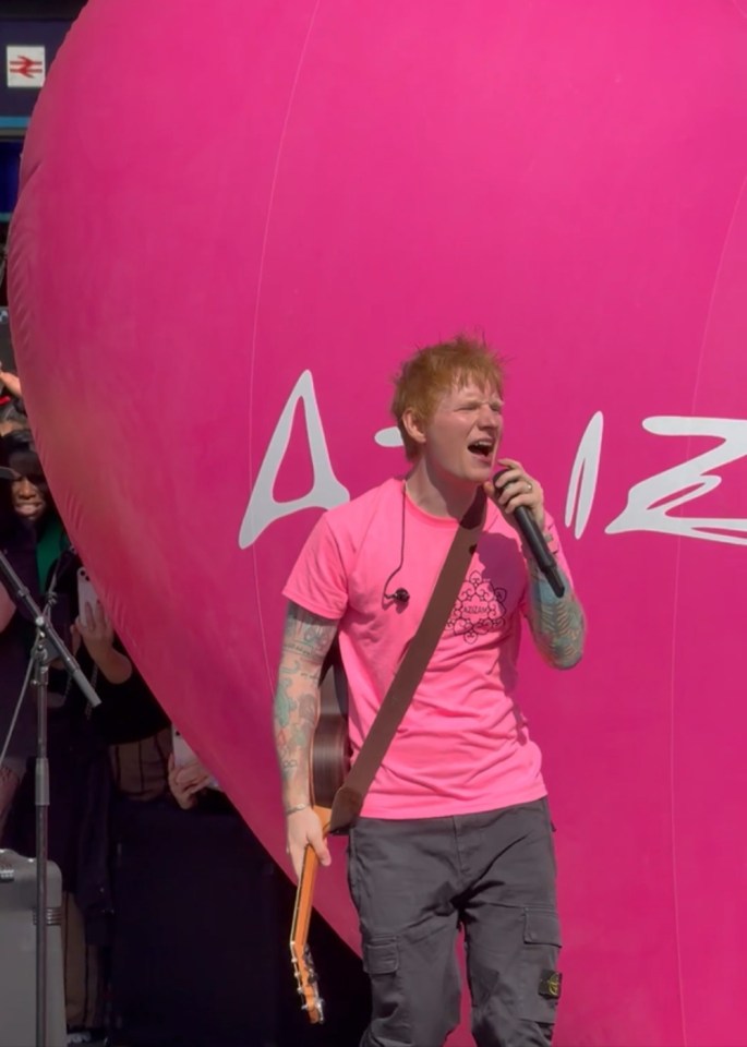 Ed Sheeran performing at King's Cross station in London, wearing a pink shirt and holding a guitar.
