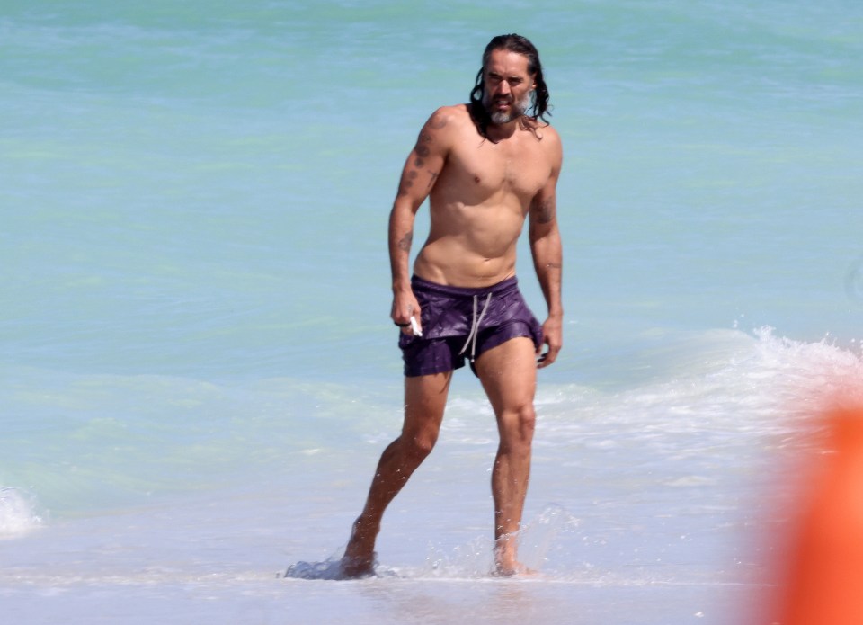 Russell Brand walking in the ocean wearing purple swim trunks.
