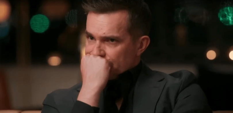 A man in a suit sits and holds his fist to his mouth, appearing upset.