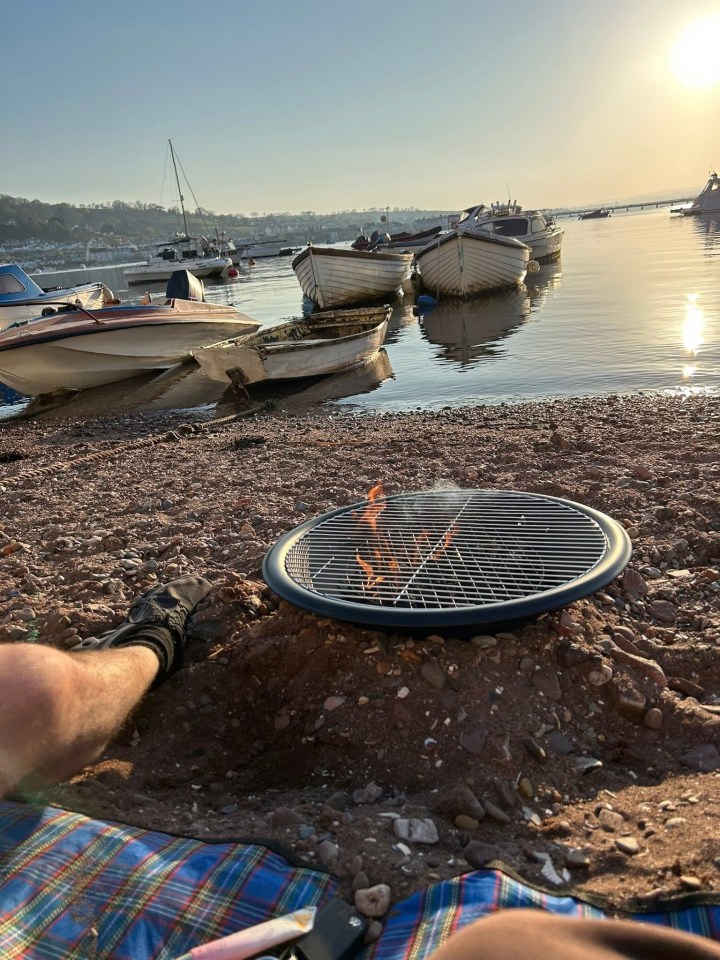 Portable fire pit burning on a beach with boats in the background.