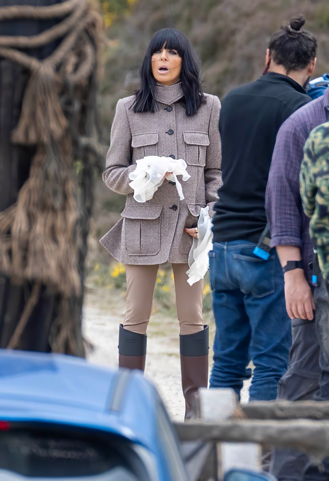 A woman in a beige jacket and brown boots stands on a film set.