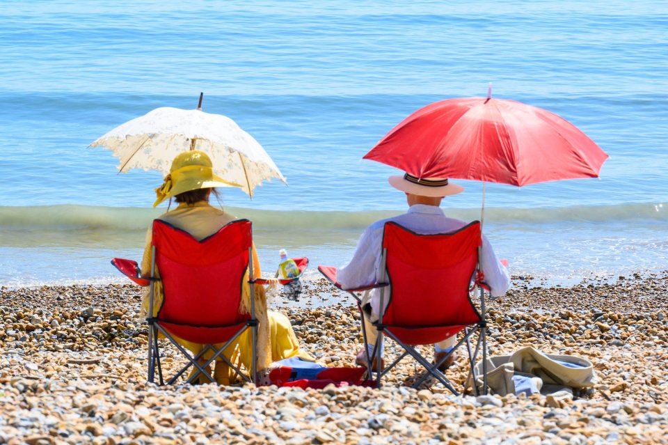 Two people sitting in chairs on a pebble beach, each under a parasol, facing the sea.