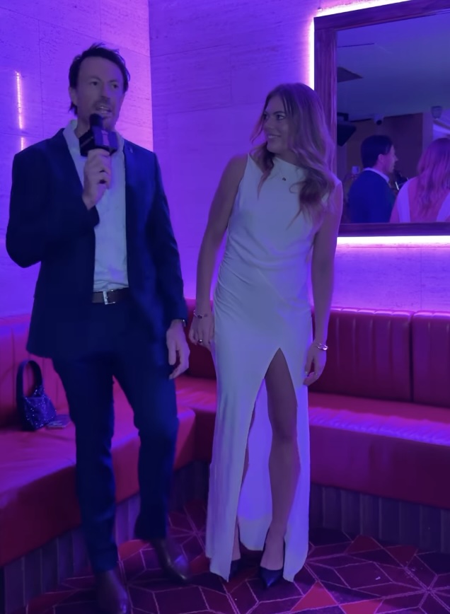 A man in a suit holding a microphone interviews a woman in a white dress.