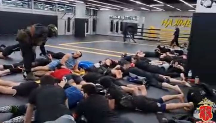 Men lying on the floor of a gym being apprehended by a person in tactical gear.