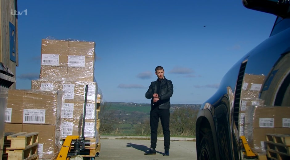 Emmerdale's Caleb looking at his watch near stacked boxes and a car.