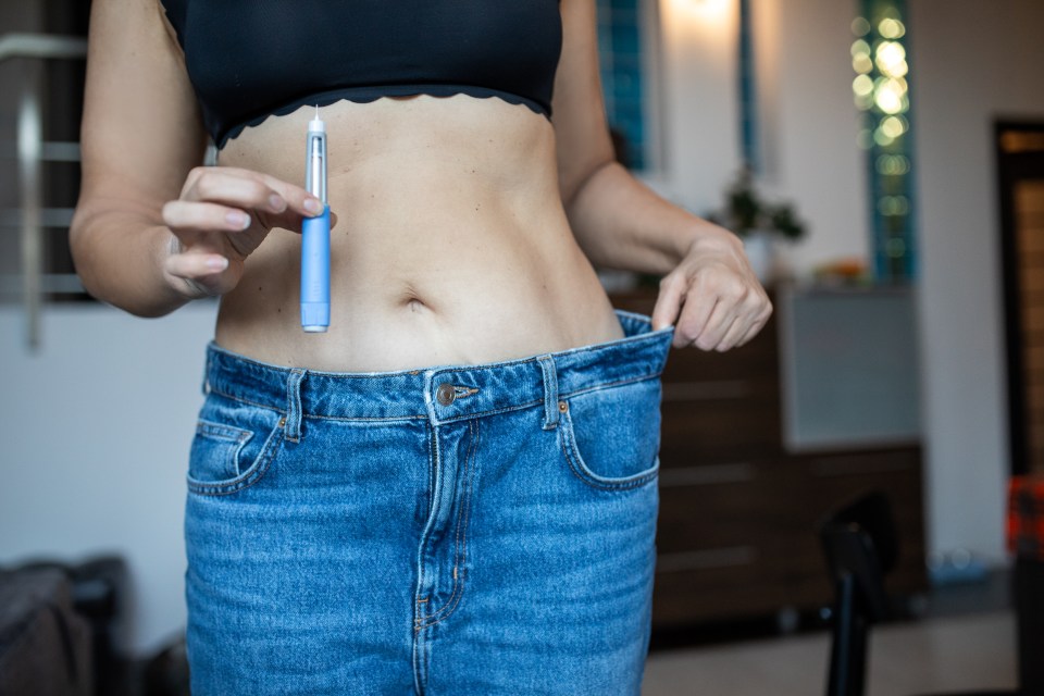 Woman holding a semaglutide injector and showing loose jeans after weight loss.