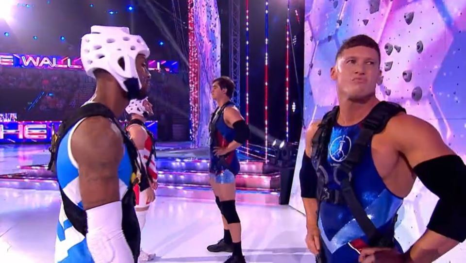 Contestants on a game show stand before a climbing wall.