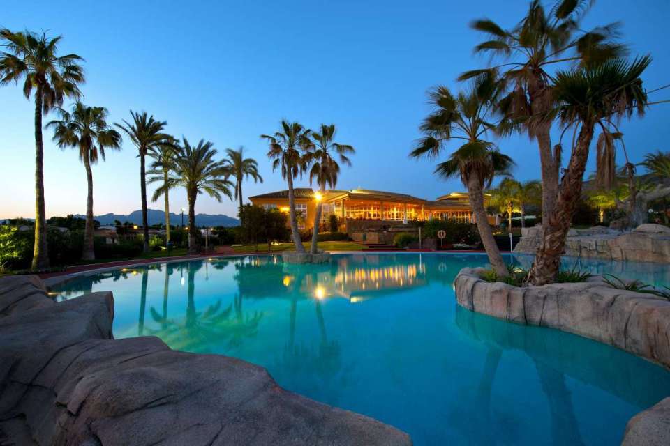 Swimming pool at night, illuminated building in the background, palm trees.