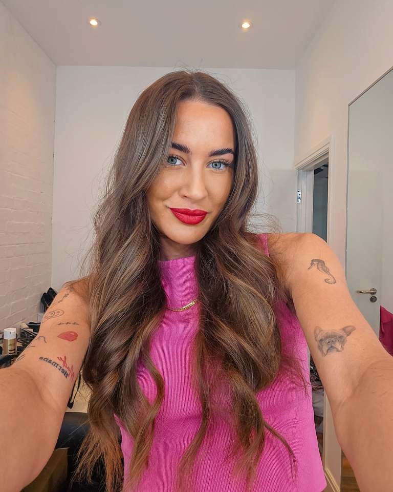 Woman with long brown hair and tattoos, wearing a pink top.
