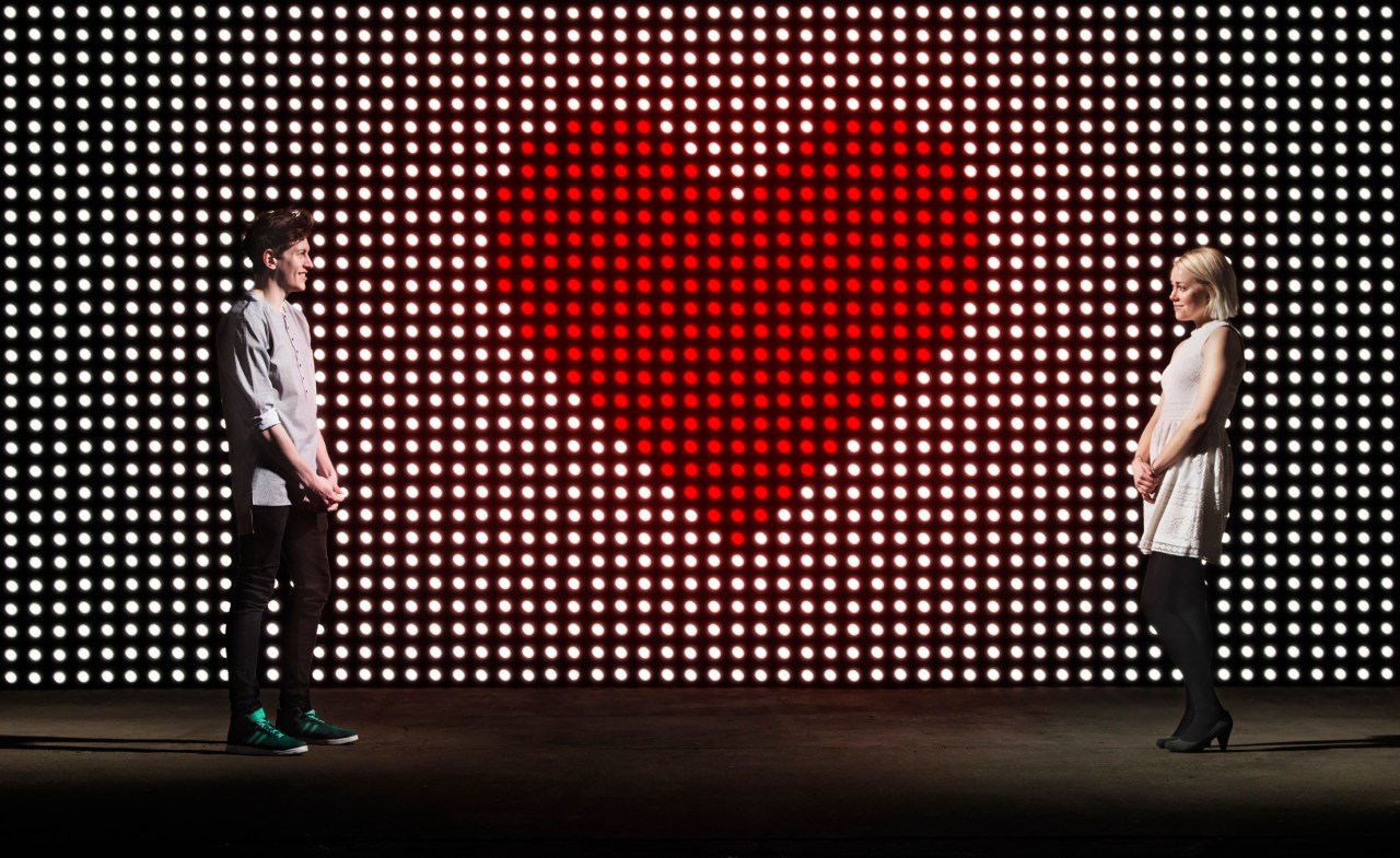 Young couple standing on either side of a light wall with a large heart shape in the center.