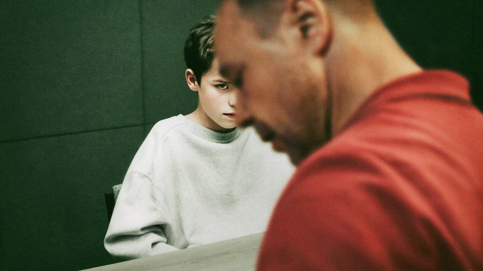 A thirteen-year-old boy sits across from a man, both appearing serious.