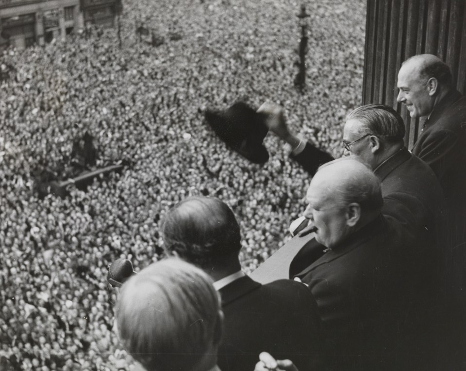 Black and white photo of Winston Churchill and other members of the cabinet addressing a large crowd from a balcony on VE Day.